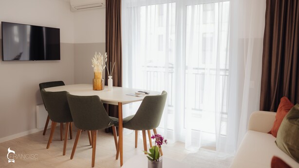 A modern living room with a dining area, a flat-screen TV mounted on the wall, and a large window with sheer curtains. - ZEN Garden Apartments (Cluj-Napoca)