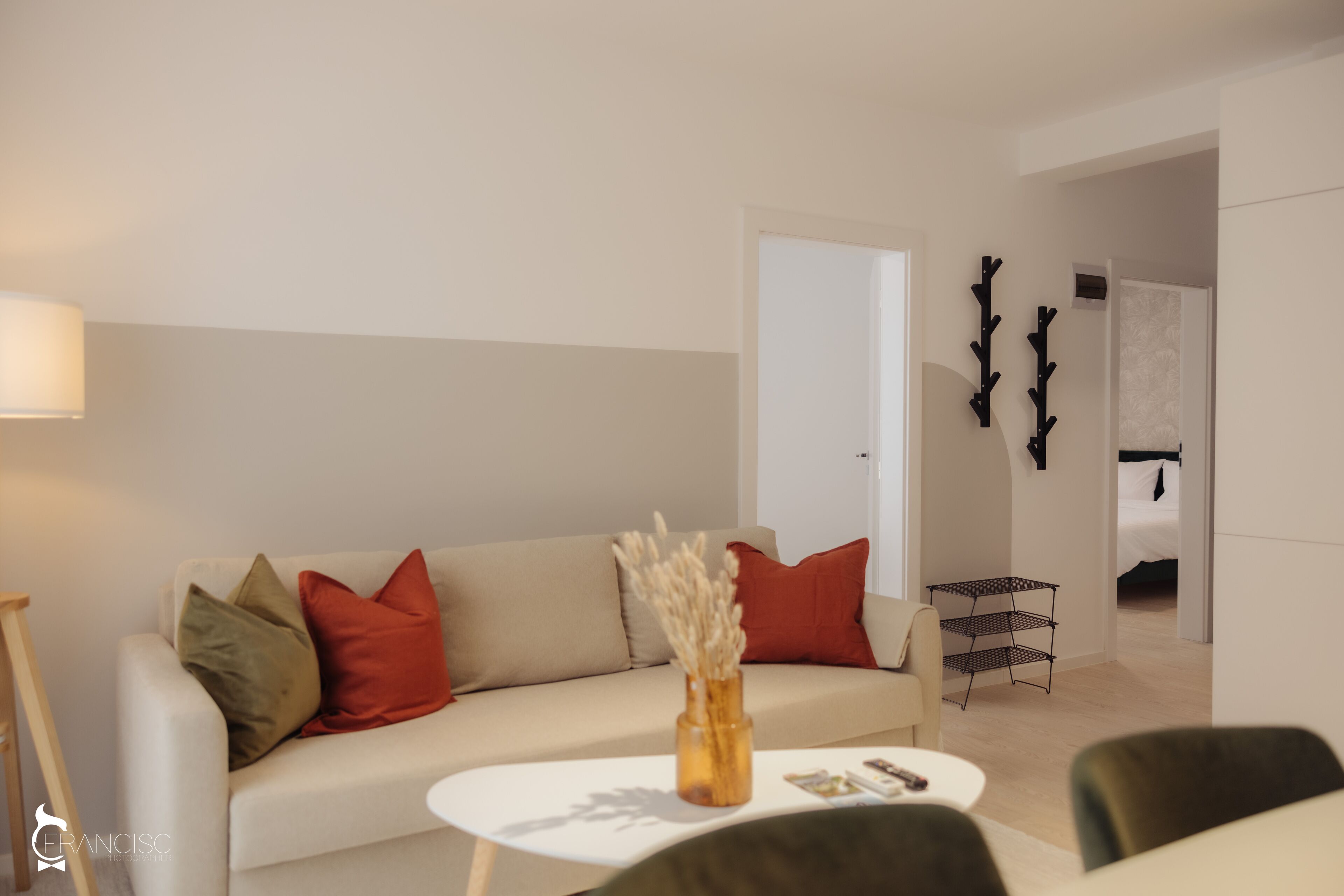 A modern living room with a white sofa, red and green cushions, a small white table, and a vase with dried plants.