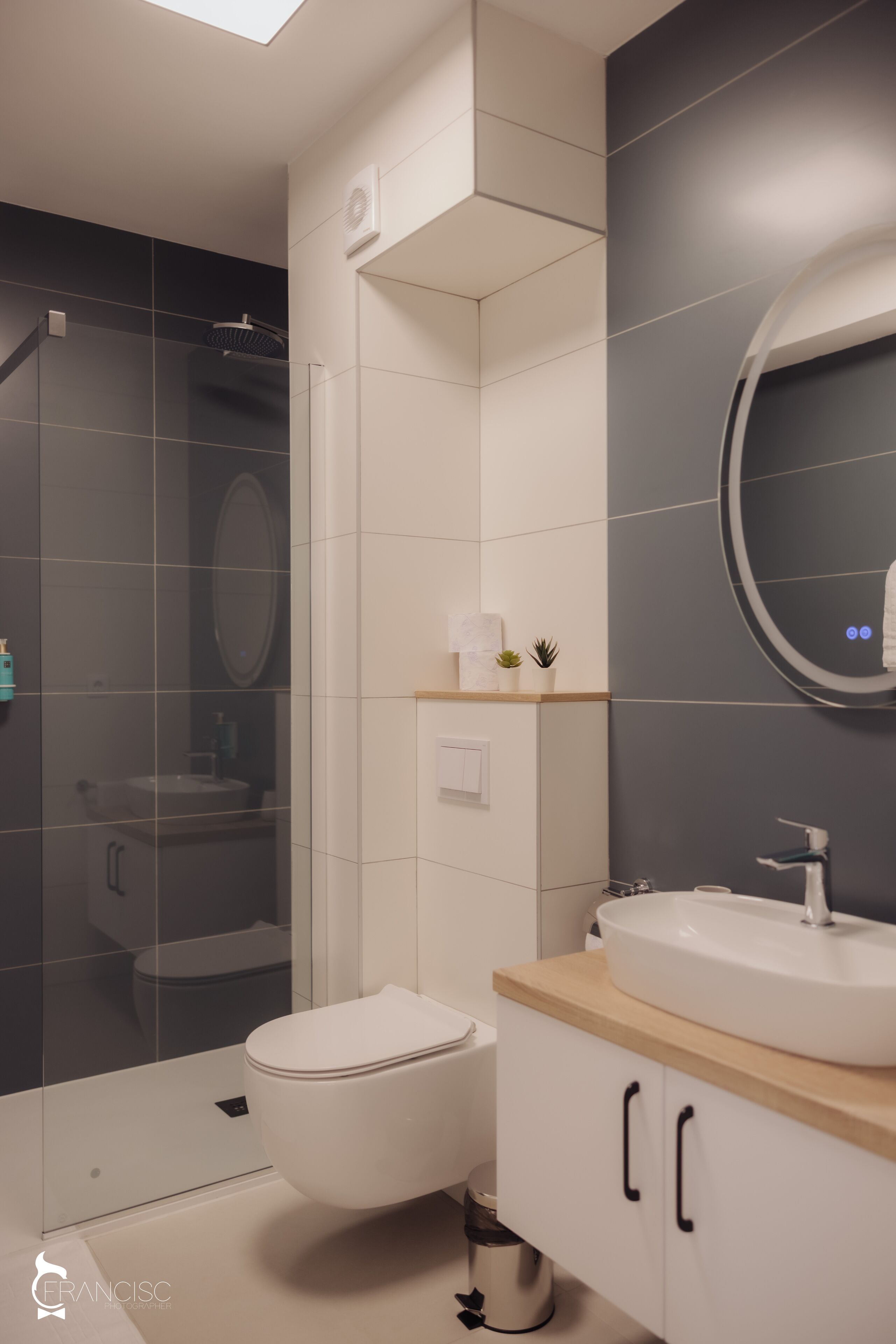 A modern bathroom with a glass shower enclosure, a white toilet, a sink with a wooden countertop, and a mirror with a circular frame.