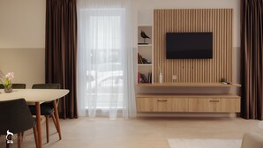 A modern living room with a dining area, a flat-screen TV mounted on a wooden panel, and a large window with sheer curtains. - ZEN Garden Apartments (Cluj-Napoca)