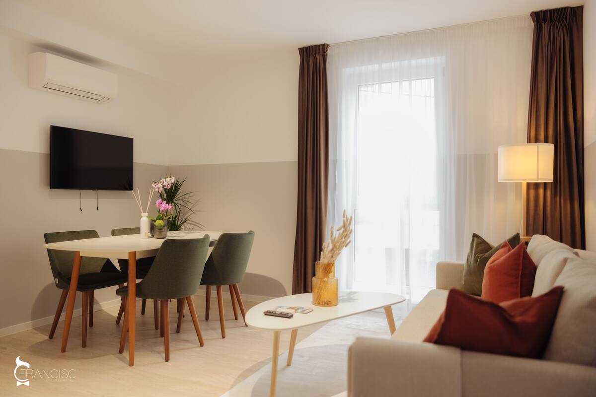 Ein modernes Wohnzimmer mit einem an der Wand befestigten Flachbildfernseher, einem Essbereich mit einem weißen Tisch und grünen Stühlen sowie einer Couch mit vielen Kissen.