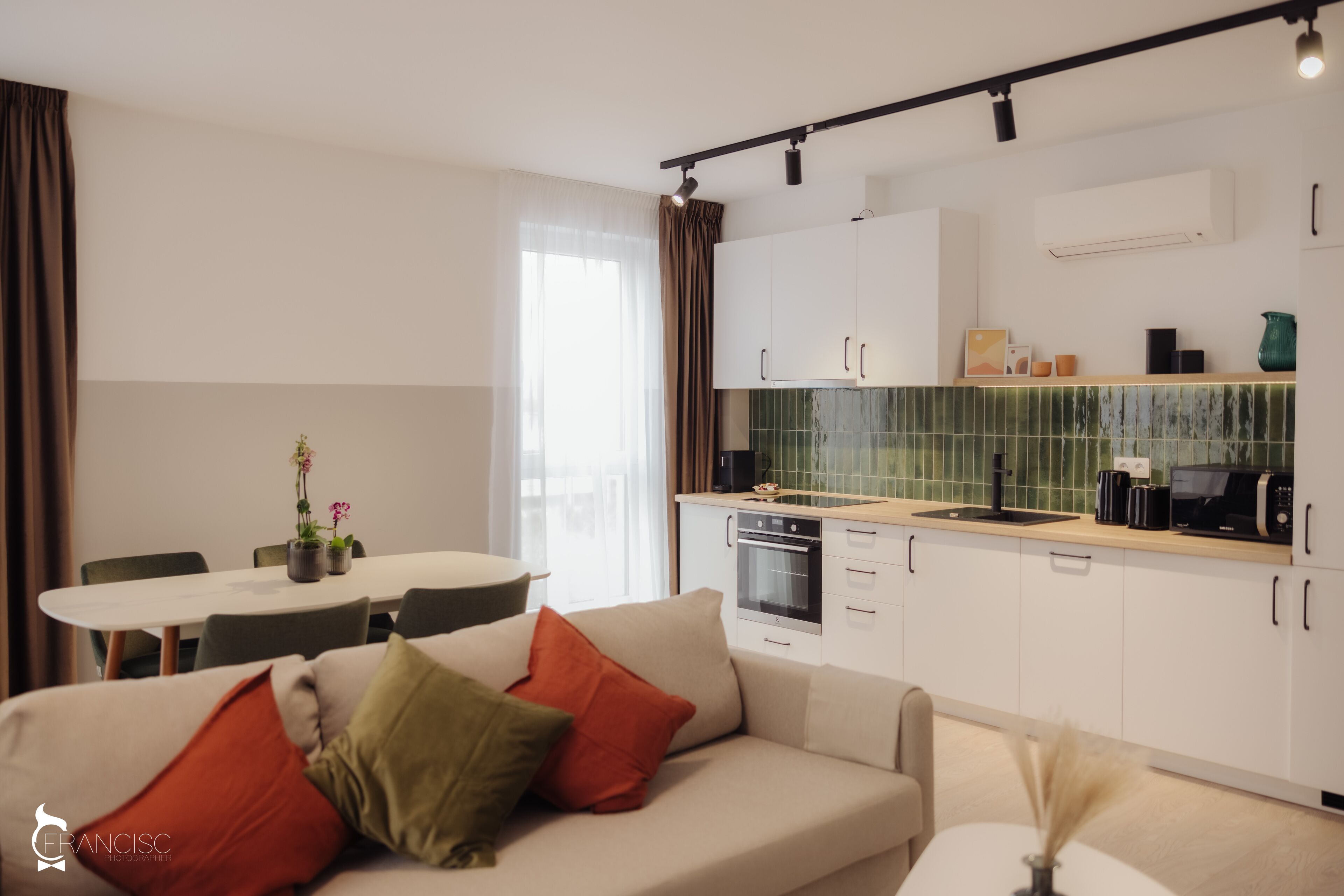A modern kitchen with white cabinets, a black oven, and a green backsplash. There is a dining table with chairs and a sofa with red and green cushions.