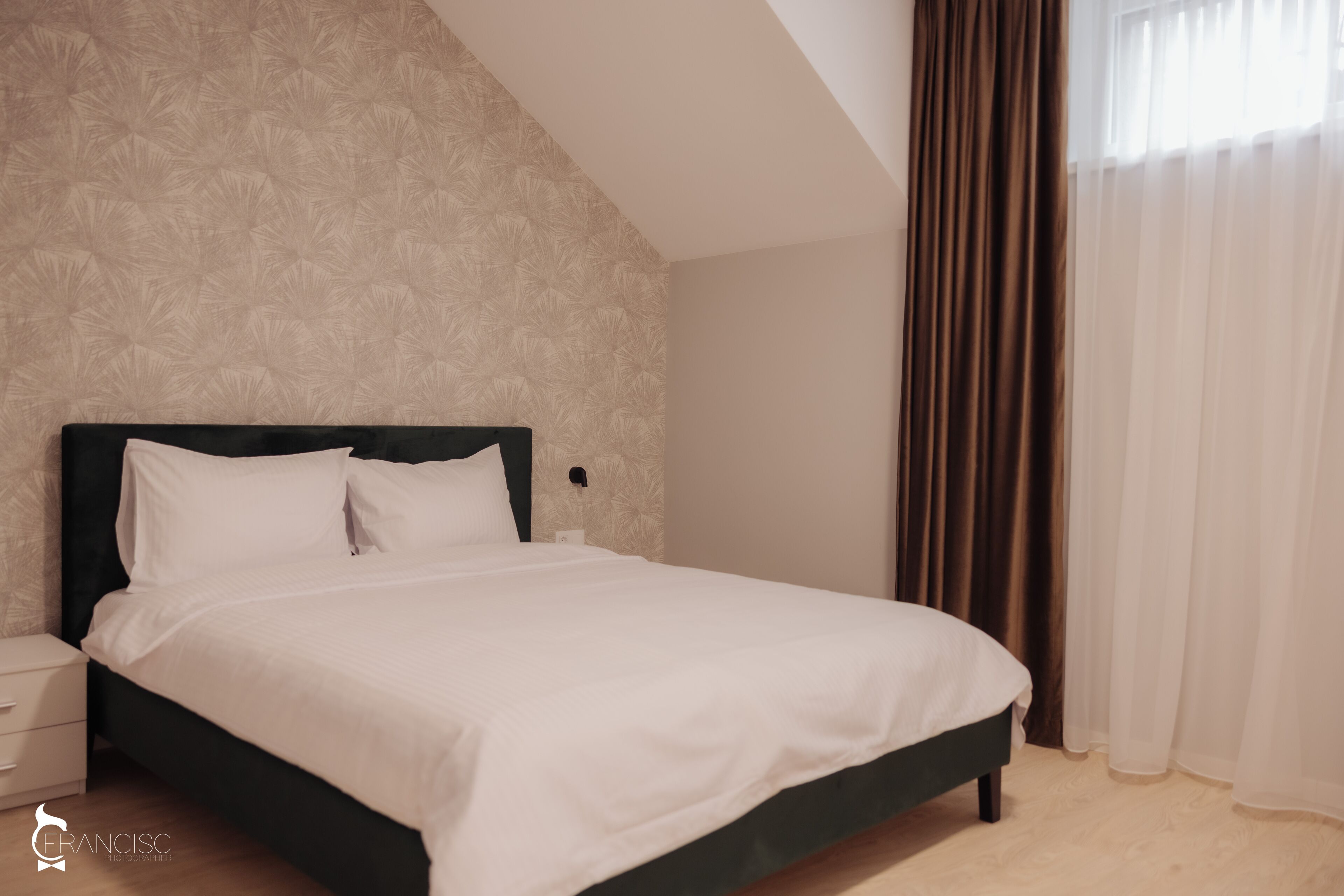 A neatly made bed in a room with a patterned wallpaper, a nightstand, and a window with curtains.