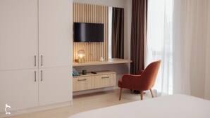 A modern hotel room with a flat-screen TV mounted on a wooden panel wall, a wooden desk with a lamp, a red chair, and a large window with curtains. - ZEN Garden Apartments (Cluj-Napoca)