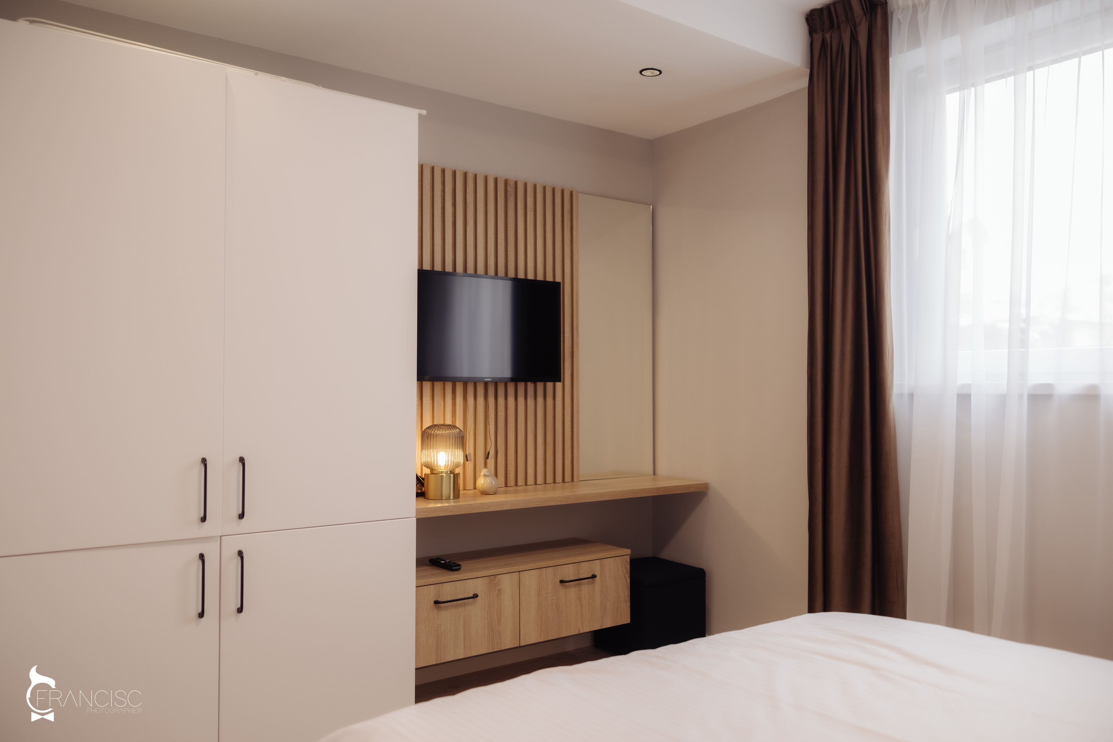 A hotel room with a bed, a TV mounted on a wooden panel wall, a desk with a lamp, and a wardrobe.