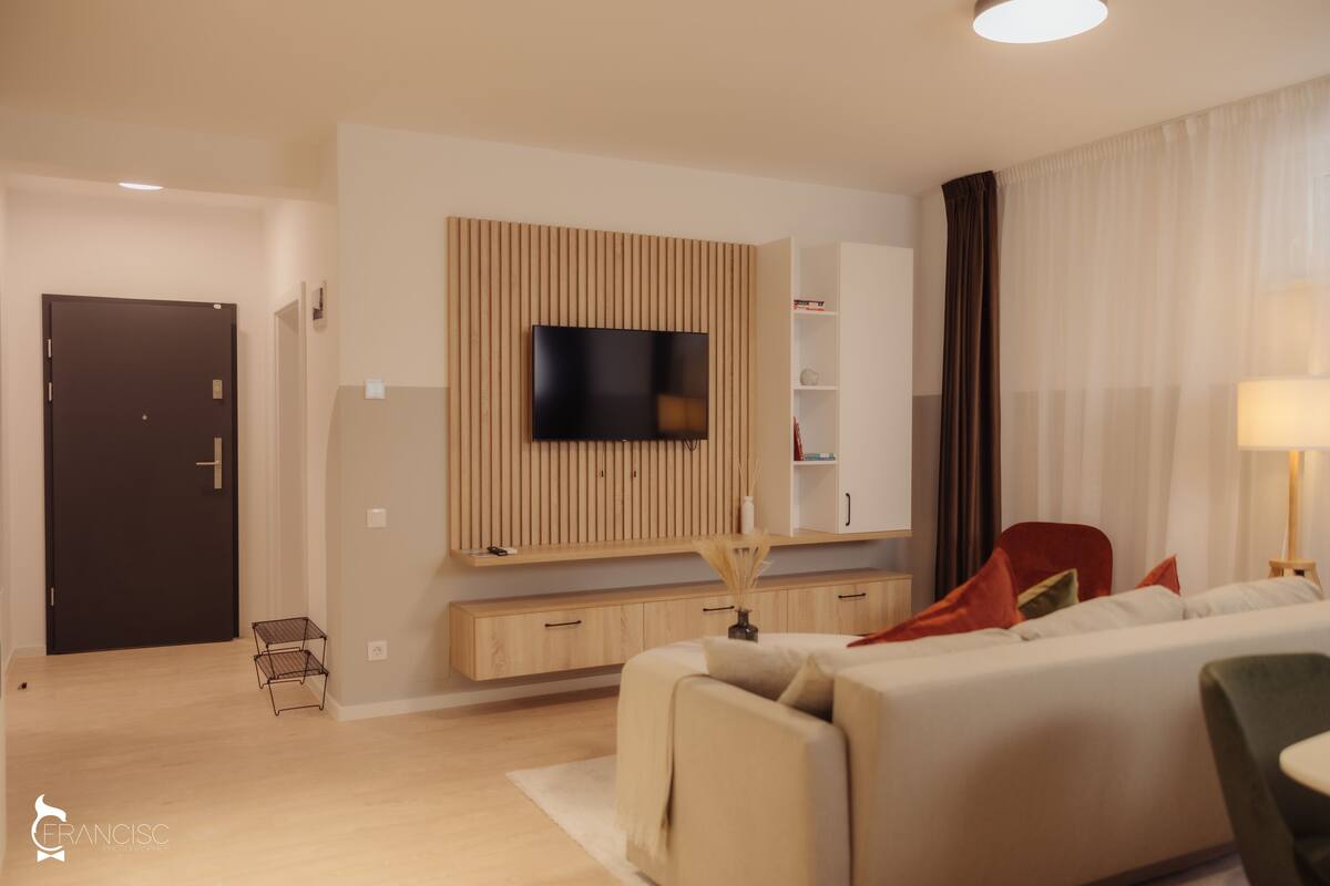 Ein modernes Wohnzimmer mit einem Flachbildfernseher an einer Holzvertäfelung, einer Couch, einem roten Sessel und einem kleinen Beistelltisch mit einer Lampe.