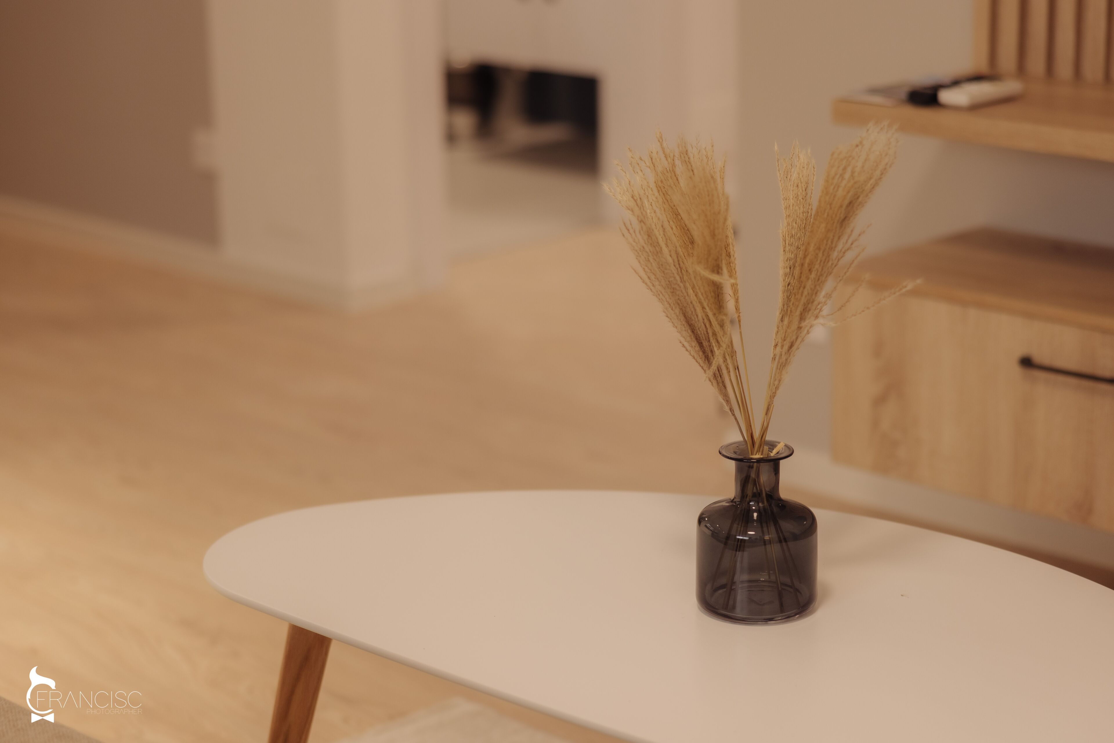 Una mesa blanca con un florero de vidrio oscuro que contiene plantas secas.