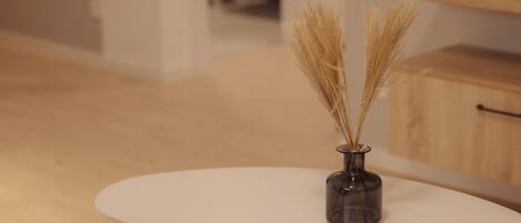 Una mesa blanca con un florero de vidrio oscuro que contiene plantas secas.