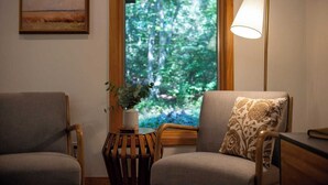 Une pièce comprenant deux fauteuils, une lampe sur pied, un tableau et une fenêtre donnant sur une forêt.