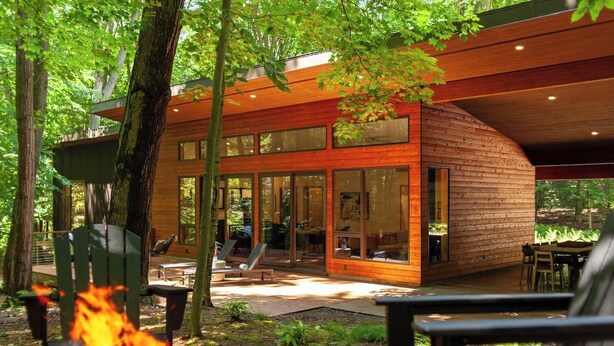 Une cabane moderne en bois, dotée de grandes fenêtres, d’une terrasse et d’un foyer extérieur.