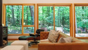 Un salon chaleureux avec de grandes fenêtres donnant sur la forêt, une cheminée et des fauteuils confortables.