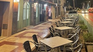 Un café al aire libre con mesas y sillas, un edificio con balcón y una palmera al fondo.