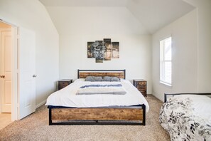 A bedroom with a wooden bed frame, a white bedspread, and a gray and white floral comforter. There is a wall art piece above the bed and a window with blinds on the right.