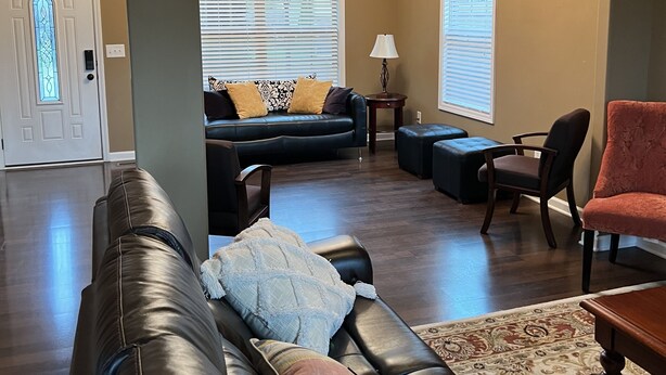A spacious living room with a dark brown leather sofa, a patterned ottoman, and a red chair. A chandelier hangs from the ceiling, and a large window with blinds is visible. - Five Stars Luxury Clean Home (Clarksville)