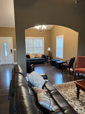 A spacious living room with a dark brown leather sofa, a patterned ottoman, and a red chair. A chandelier hangs from the ceiling, and a large window with blinds is visible.