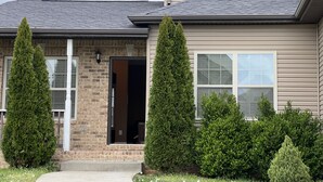 A two-story house with a brick facade, a dark roof, and a front door open. There are two tall, conical trees in front of the house. - Five Stars Luxury Clean Home (Clarksville)