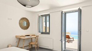 Un dormitorio con un escritorio de madera, una silla, una cama y vistas al mar a través de una puerta abierta.