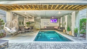 A covered outdoor swimming pool area with seating and a TV screen displaying 'Hilton Head Island'. - (119) Heated Pool, Hot Tub, Walk to the Beach, Elevator, EV Charger (Hilton Head Island)