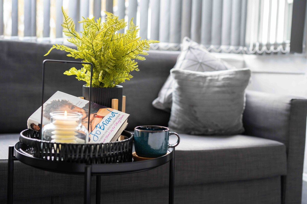 Una sala de estar con un sofá gris, una mesita negra con una planta, una vela, una taza y una revista.