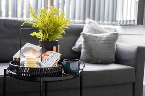 A living room with a grey sofa, a small black side table with a plant, a candle, a mug, and a magazine.