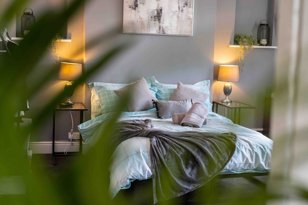 A neatly made bed with a blue and white checkered duvet, two pillows, and a blanket. There are two bedside lamps on either side of the bed.