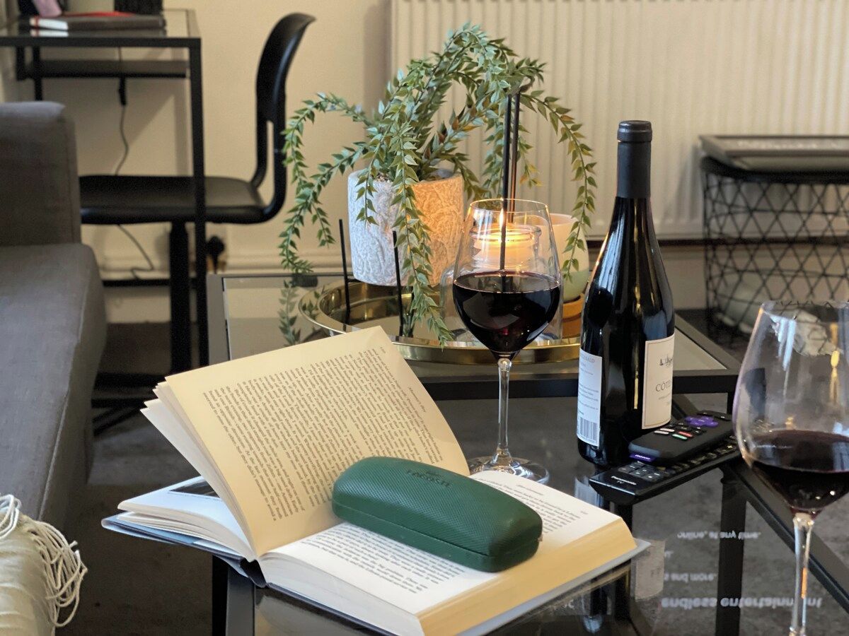 Een boek, een groen kussen, een wijnfles en glazen op een glazen tafel, met een plant en kaars op de achtergrond.