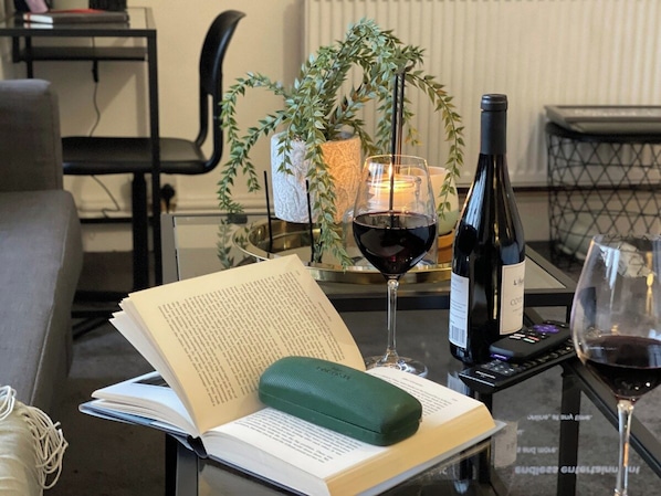 A book, a green cushion, a wine bottle, and glasses on a glass table with a plant and candle in the background.