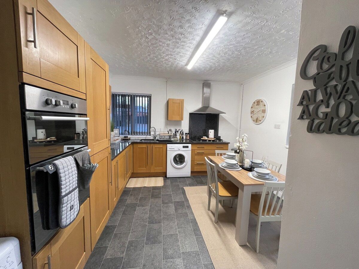 Una cucina compatta con mobili in legno, forno a incasso, lavatrice e zona pranzo con tavolo e sedie.