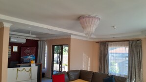 A living room with a sofa, a coffee table, and a chandelier. There is a door leading to another room and a window with curtains. - Ramatie Guest Palace (Kasane)