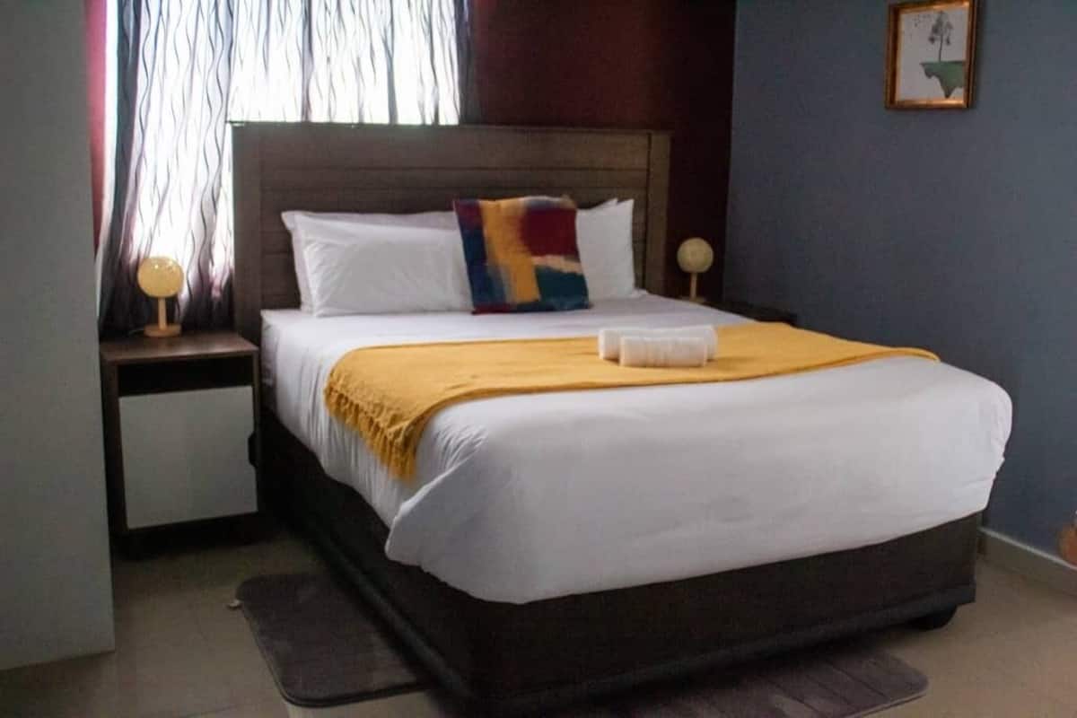 A neatly made bed with white linens and a yellow throw blanket, a wooden headboard, a bedside lamp, and a framed picture on the wall.