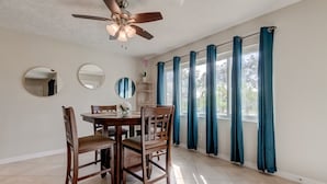 A dining area with a wooden table and chairs, a ceiling fan, and large windows with blue curtains. -  Few Mins to Madeira Beach, Comfortable, Dog Friendly (Seminole)