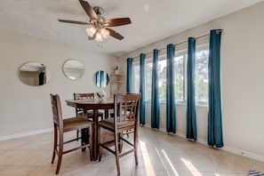 A dining area with a wooden table and chairs, a ceiling fan, and large windows with blue curtains.