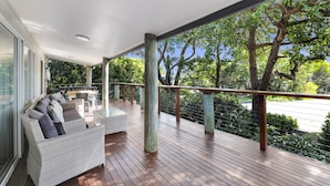 A covered outdoor area with a wooden deck, a white wicker sofa with cushions, and a glass table.