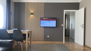 A modern living room with a flat-screen TV mounted on the wall, a wooden dining table with chairs, a grey sofa, and a wooden coffee table.