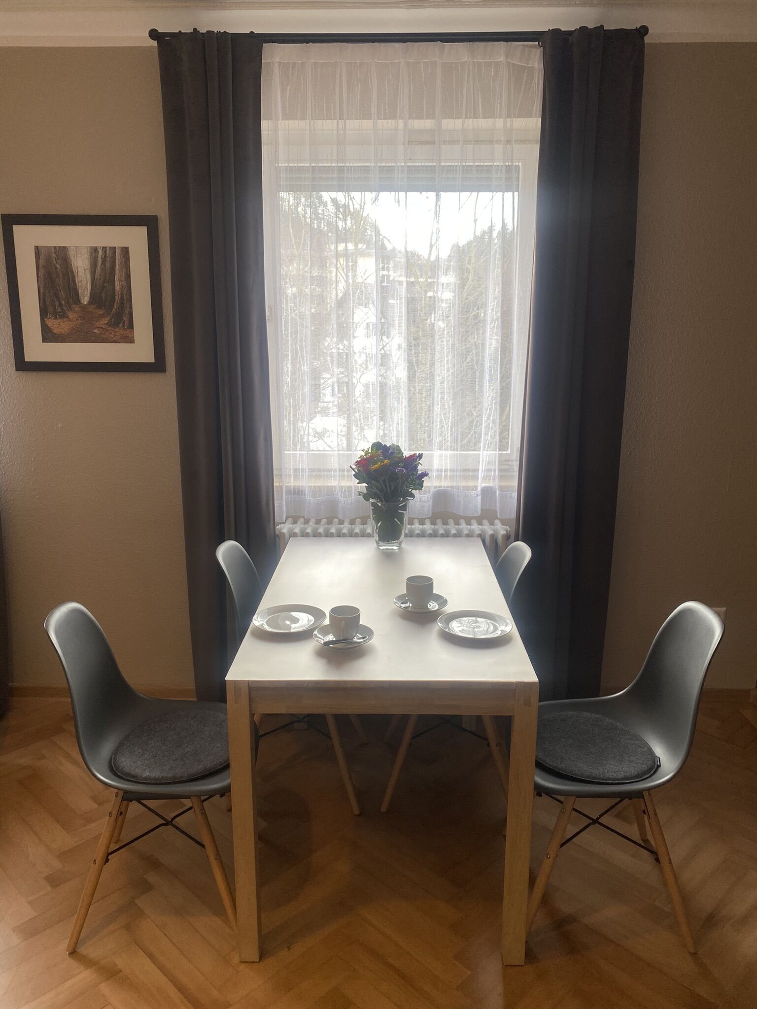 Een eethoek met een tafel gedekt voor twee personen, een vaas met bloemen en een lijst met een foto aan de muur.