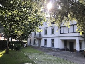 Un edificio de dos plantas con balcón, un camino de entrada pavimentado y un césped con una silla.