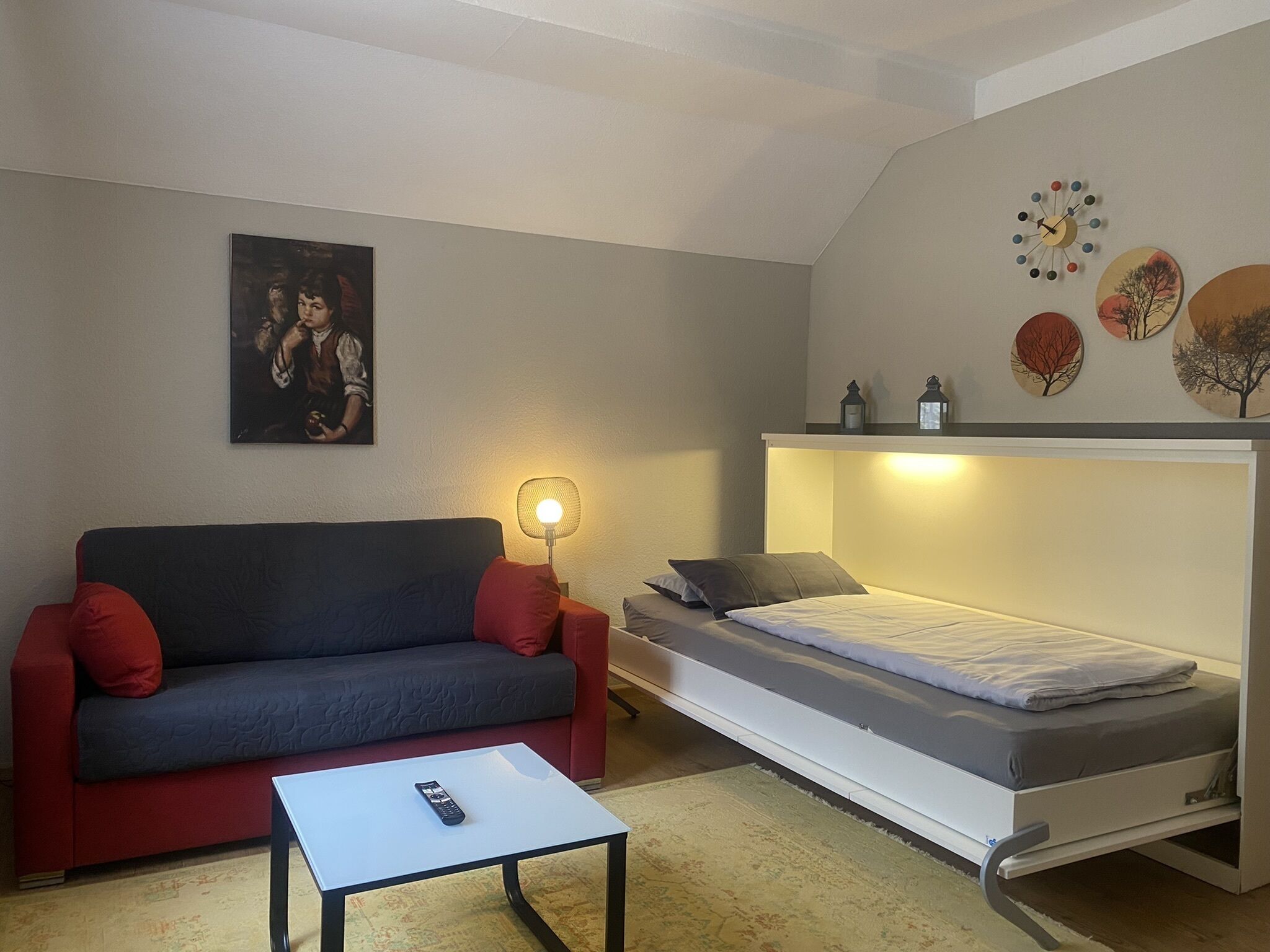A compact room with a sofa, a small table, a bed with storage, a wall clock, and a painting.