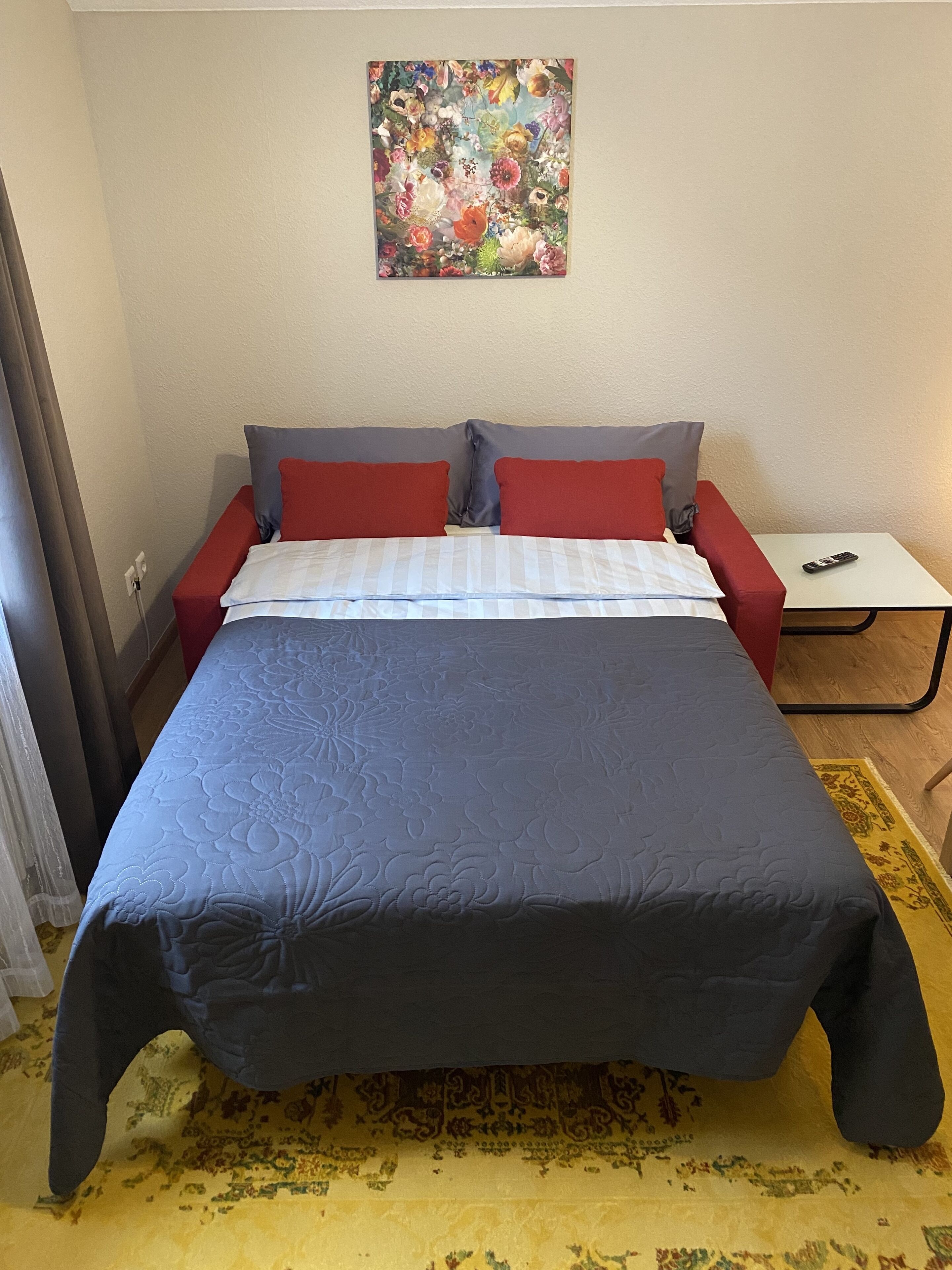 A bed with a blue and gray quilt, red and gray pillows, and a small table with a remote control beside it.
