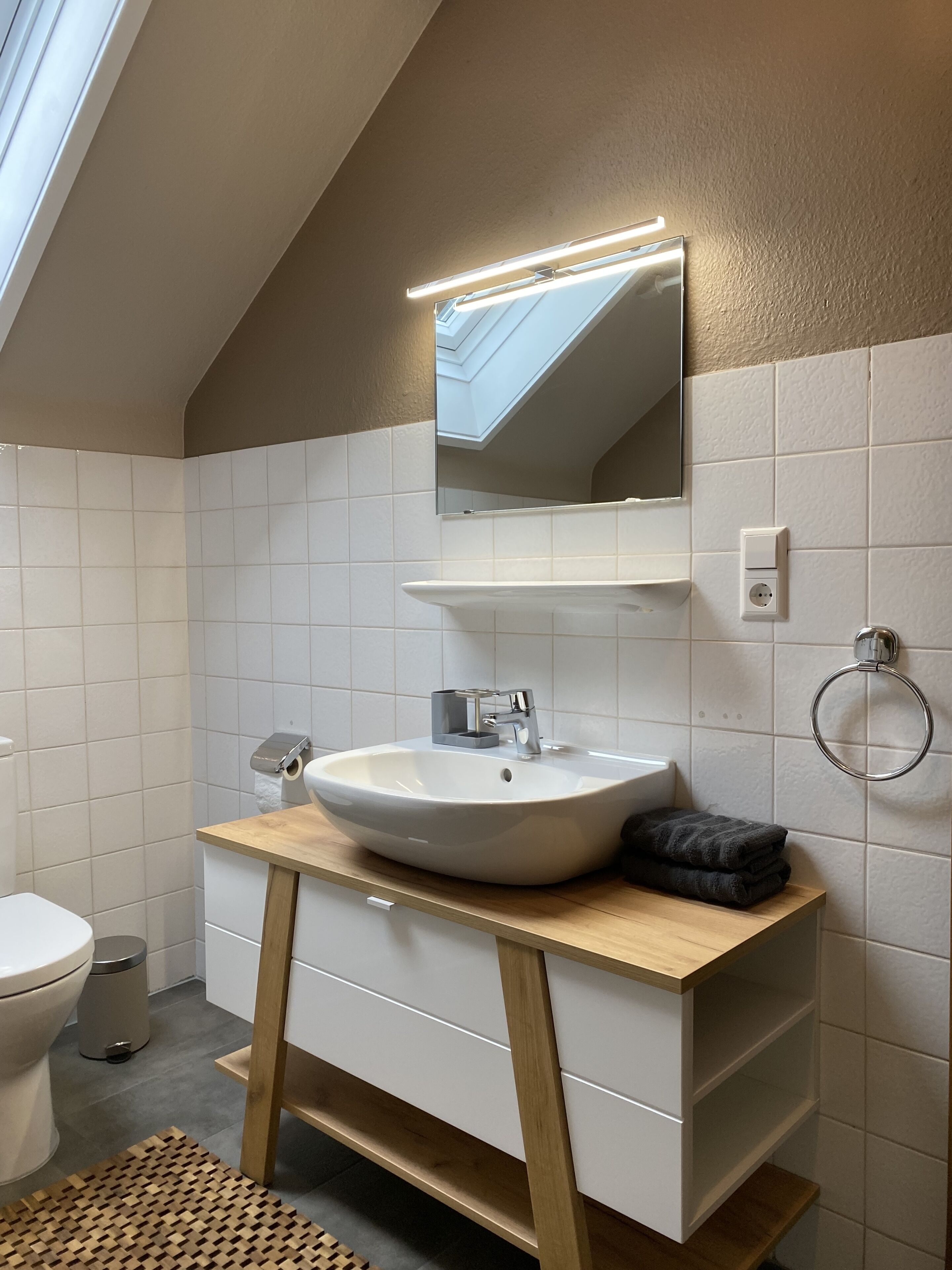 Ein modernes Badezimmer mit Dachfenster, weißen Fliesen, einem hölzernen Waschtisch, einer weißen Spüle und einem Spiegel.