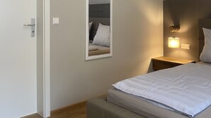 A bedroom with a bed, a bedside table with a lamp, a mirror on the wall, and a door.