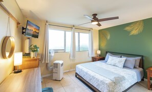 Ein Hotelzimmer mit einem Bett, einem Schreibtisch mit Lampe, einem Fernseher, einem Fenster mit Vorhängen, einem Deckenventilator und einer Wand mit Palmenblatt-Motiv.