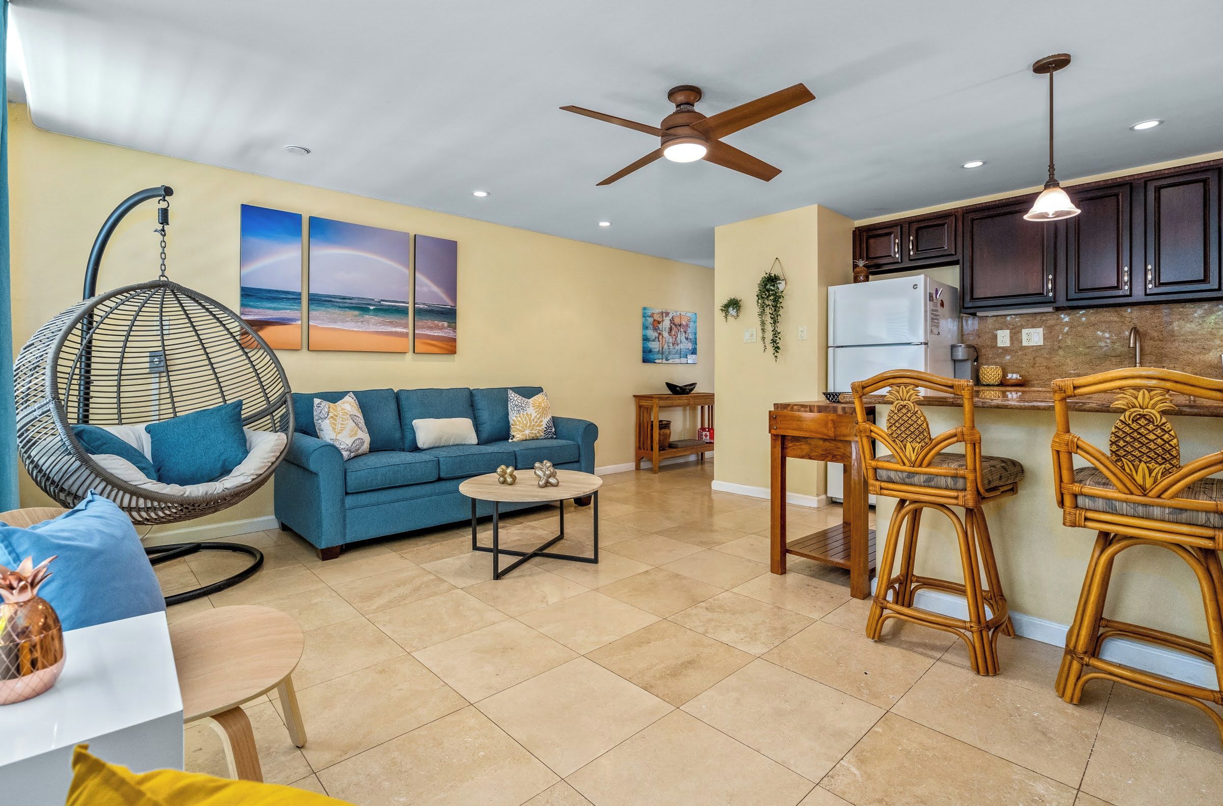 A living room with a blue sofa, a hanging chair, and a coffee table. There is a kitchen area with wooden cabinets and a refrigerator.