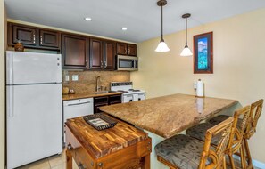 Une cuisine compacte avec des meubles de couleur bois foncé, un réfrigérateur blanc, un four à micro-ondes et une cuisinière. On y trouve une table à manger en bois avec des chaises, et une suspension lumineuse est accrochée au-dessus.