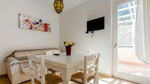 A dining area with a white table and chairs, a sofa, a painting, and a TV on the wall.