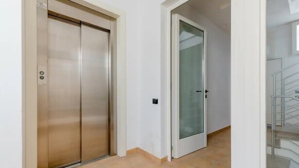 A hallway with a metallic elevator and a glass door, leading to a staircase.