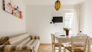 A room with a striped sofa, a dining table with chairs, a TV mounted on the wall, and a hanging light fixture.