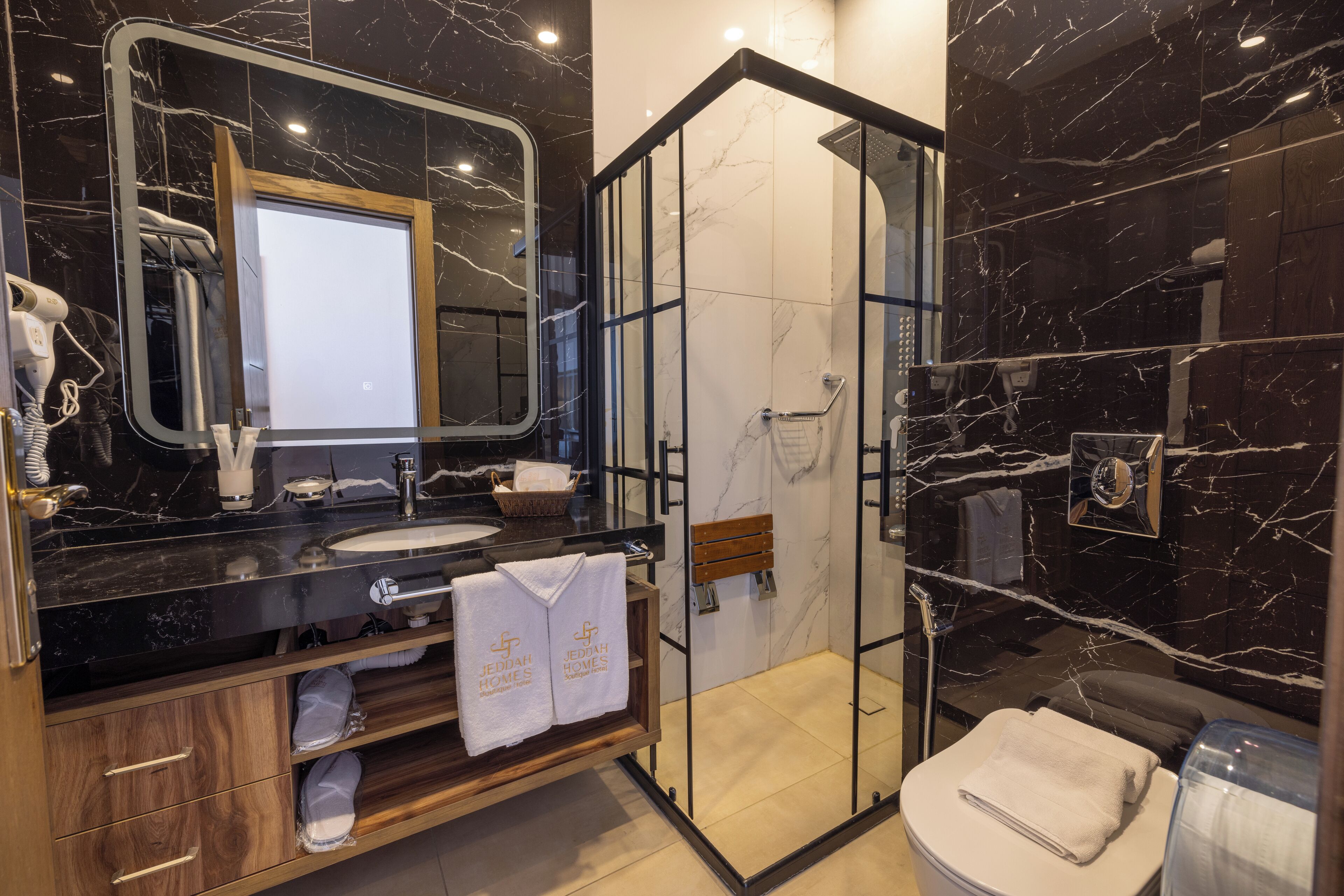 Une salle de bain moderne avec un meuble-lavabo en marbre noir, une douche à parois de verre et un meuble en bois où sont rangées les serviettes.
