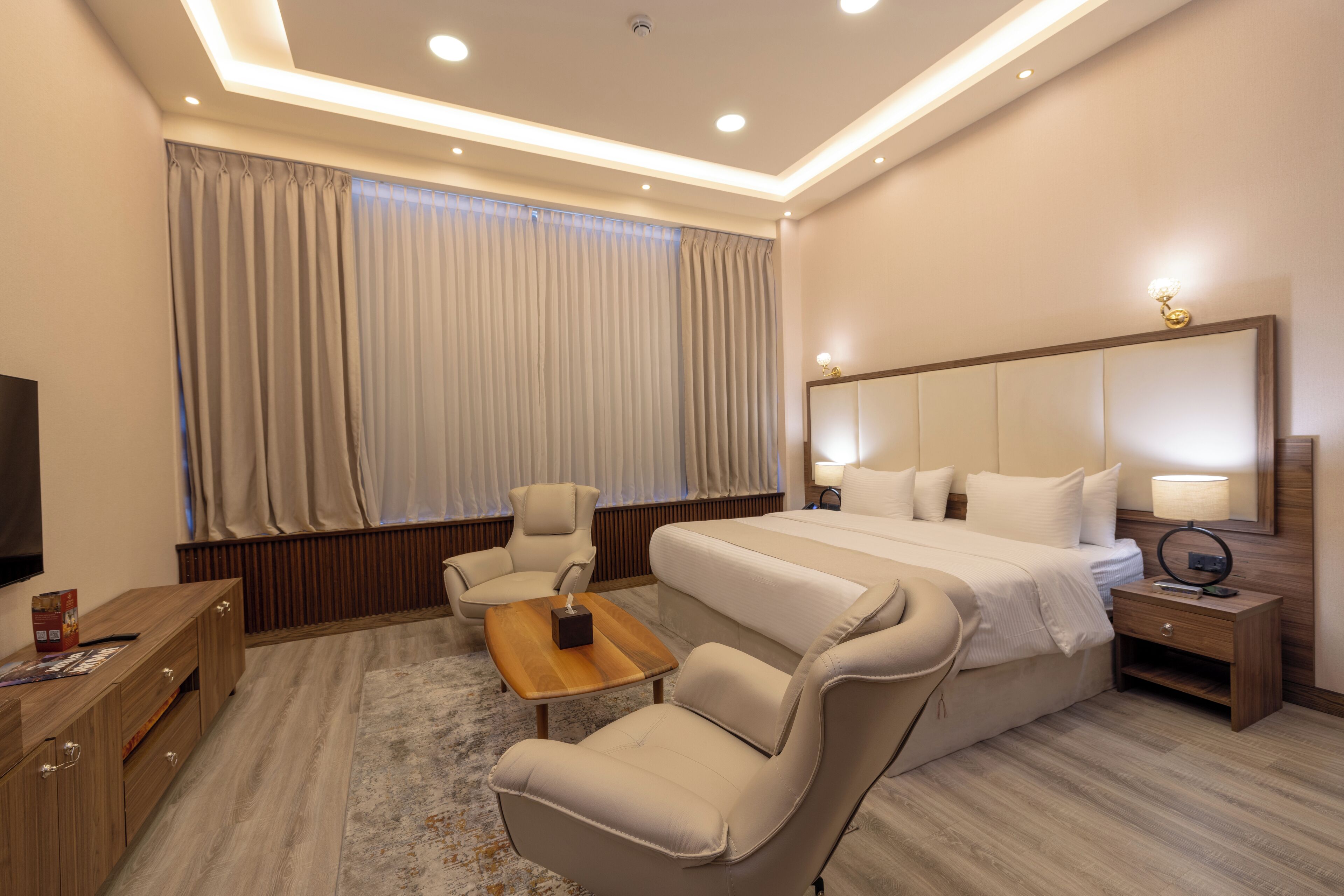 Ein modernes Hotelzimmer mit einem großen Bett, einer Sitzecke mit Stühlen, einem Holztischchen, einem Fernseher und einem großen Fenster mit Vorhängen.
