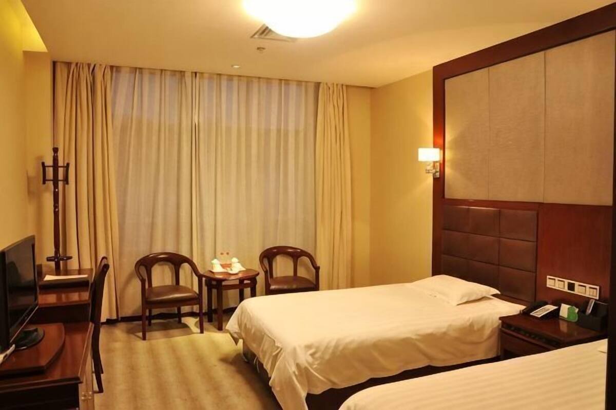 Quarto de hotel com duas camas, televisão, escrivaninha com cadeira, mesa pequena e janela com cortinas.