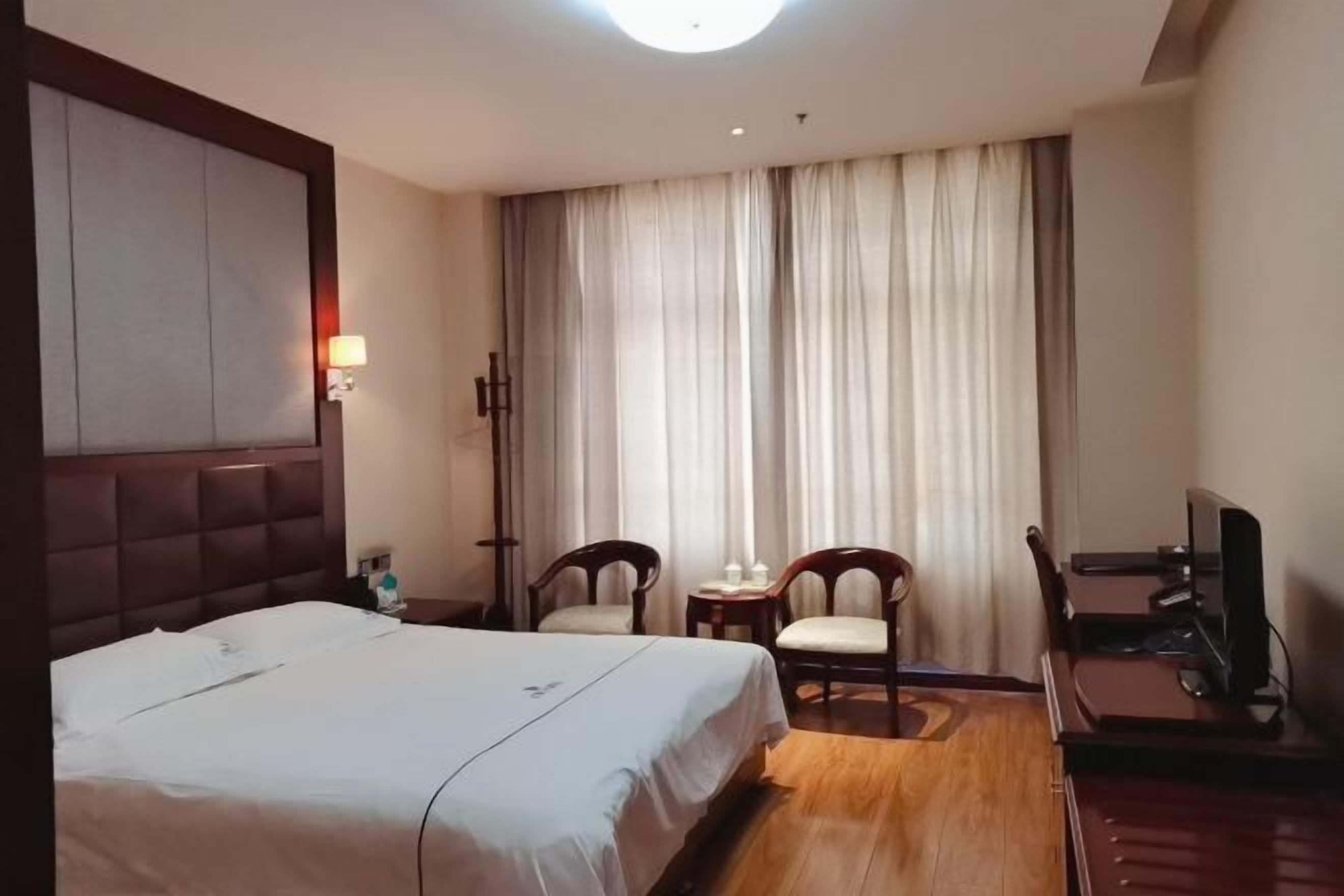 Habitación de hotel con cama, dos sillas, una mesita, televisor y un ventanal con cortinas.